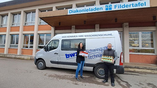 a silver grey transporter with the blue logo of company bwz Schwingungstechnik, in front of a building of the Fildertafel food bank, and in the foreground two persons holding trays with packed food items in their hands  