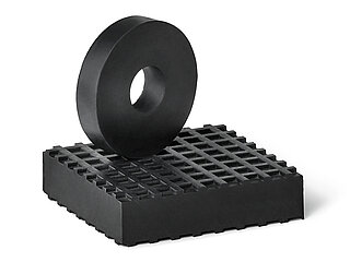 eine schwarze, quadratische Gummiplatte aus Elastomer NBR mit kleinen quadratischen Vertiefungen auf der Oberfläche als Gleitschutzprofil, darüber eine hochkant aufgestellte schwarze, runde und glatte Gummischeibe aus Elastomer NBR mit Loch in der Mitte, freigestellt auf weißem Hintergrund