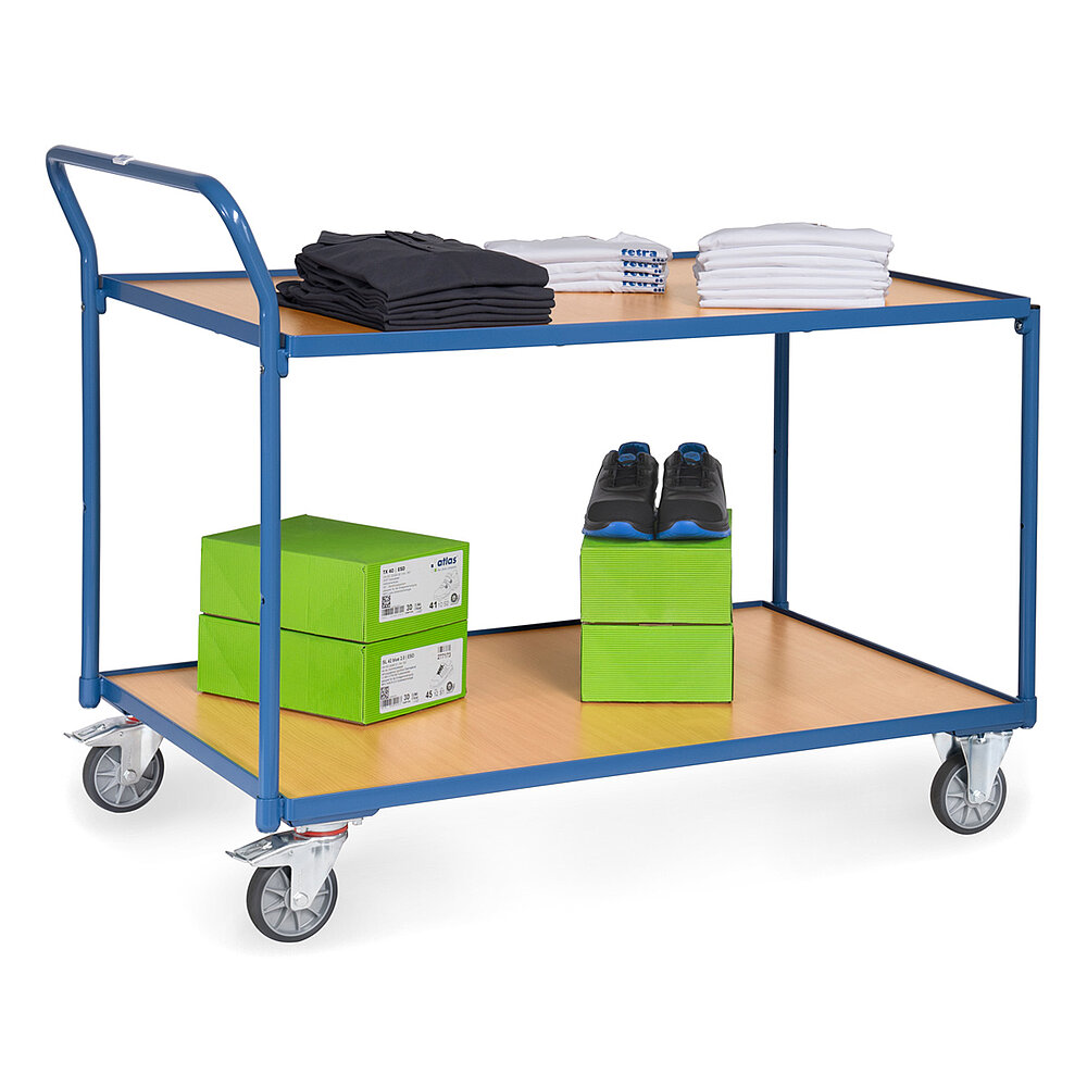ein blauer FETRA® Tischwagen aus Stahlrohr und Profilstahl mit zwei großflächigen Etagen, nach hinten gebogenem Schiebebügel, hellen Holzeinlegeböden, zwei Bockrollen vorne, zwei feststellbaren Lenkrollen hinten und beladen mit grünen Schuhkartons und T-Shirts, freigestellt auf weißem Hintergrund