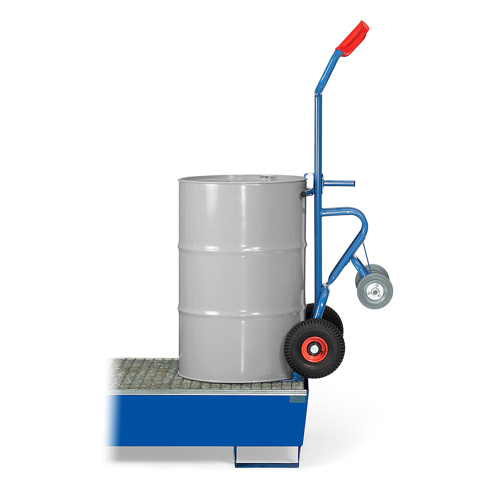 eine blaue FETRA® Karre aus Stahlrohr, mit zwei Rädern mit Luftbereifung auf roten Felgen, roten Handgriffen und geschwungener Stütze für Schräglage in Ruheposition mit zusätzlicher zweiter Achse mit zwei Stützradern, beladen mit einem grauen 200 Liter Fass aus Metall, aufrecht auf  einer Ölwanne mit Gitterrost stehend, freigestellt auf weißem Hintergrund