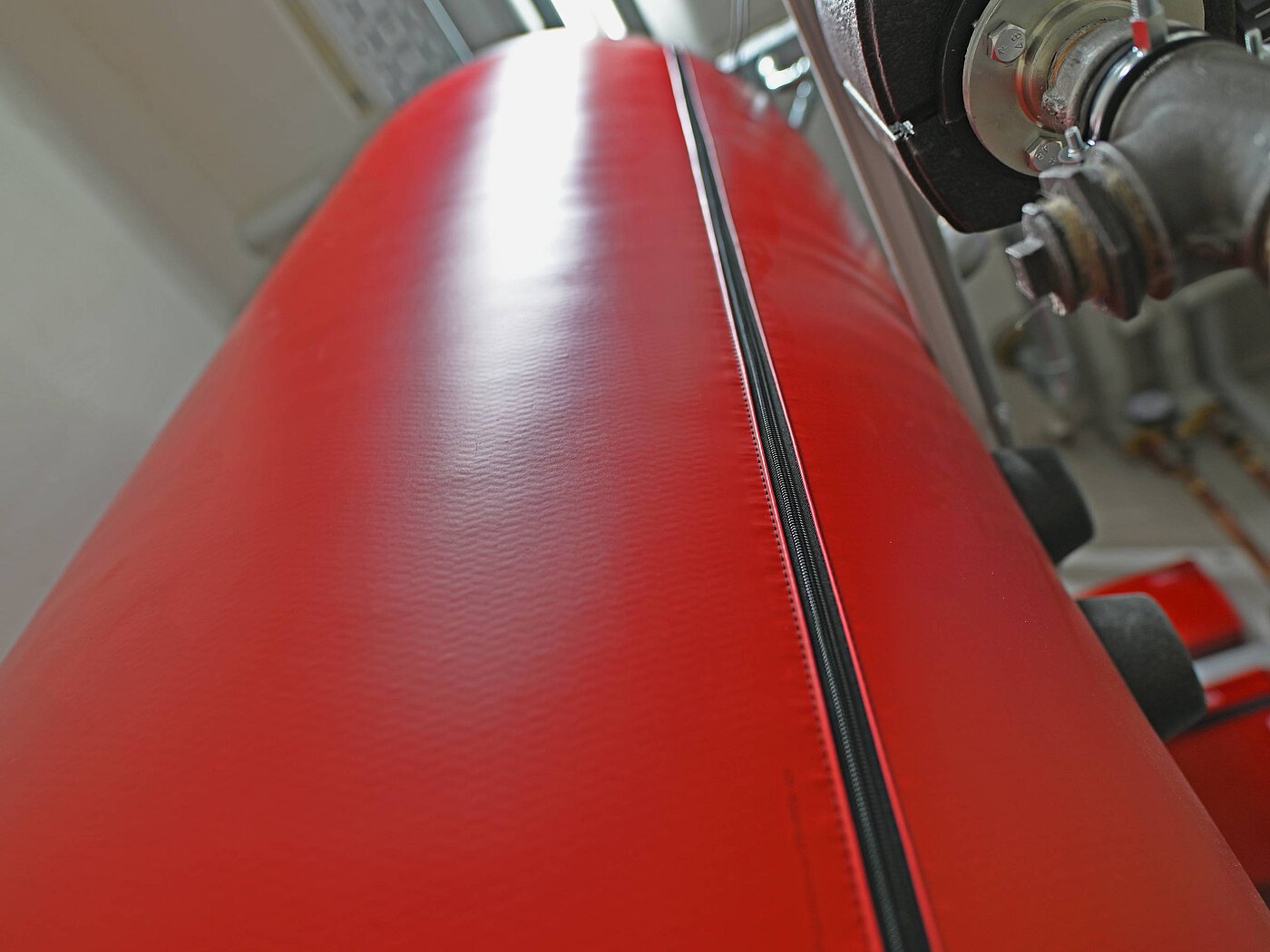 der in einer roten Isolierhülle geschütze Warmwasserspeicher einer Wärmepumpenheizanlage in einem Kellerraum mit diversen Leitungen und Installationen im Hintergrund