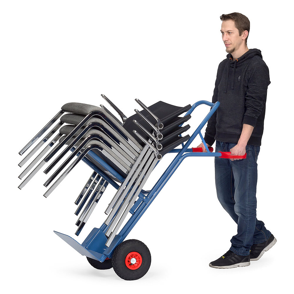 ein Arbeiter mit einer blauen, nach hinten gekippten FETRA® Karre aus Stahlrohr mit schwarzer Luftbereifung, Ladeschaufel, feststehendem, aus zwei horizontal abstehenden Holmen bestehendem Stuhltraggestell und beladen mit einem Stapel von vier Stahlrohr-Stühlen, und roten Handgriffen auf weißem Hintergrund