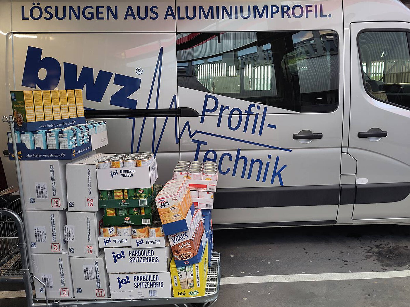 a C&C cart, fully loaded with trays and cardboard boxes of various food items such as noodles, rice, long shelf-life milk, canned fruit and breakfast cereals, parked sideways of a silver-grey delivery van with the blue logo and script of company bwz Schwingungstechnik