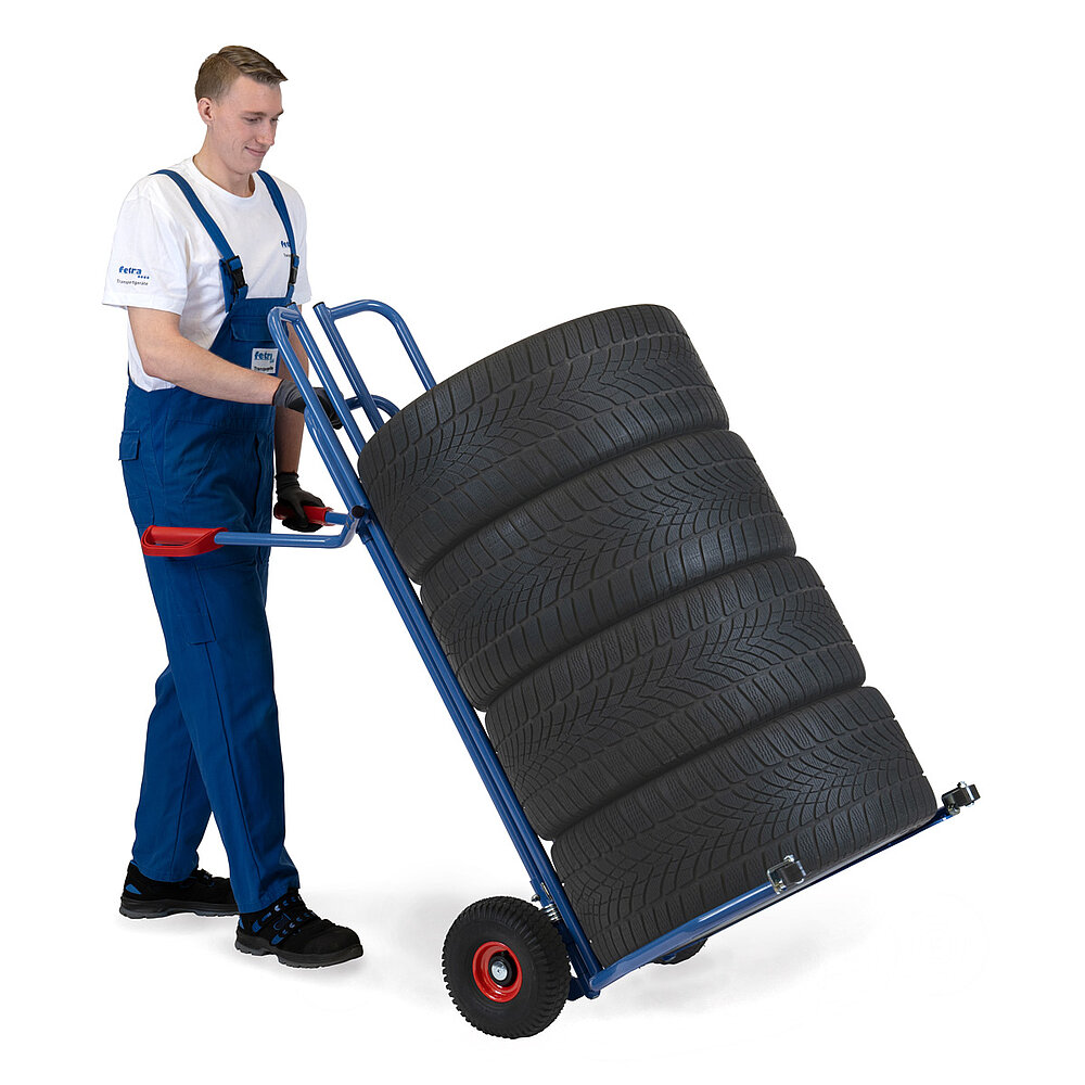ein Arbeiter hantiert mit einer nach hinten gekippten blauen FETRA® Reifenkarre aus Stahlrohr mit Luftbereifung, großen geschwungenen Spreizaufnahmebügeln mit kleinen schwarzen Kunststoffrollen am vorderen Ende und roten Handgriffen, darauf gestapelt 4 PKW-Reifen, auf weißem Hintergrund
