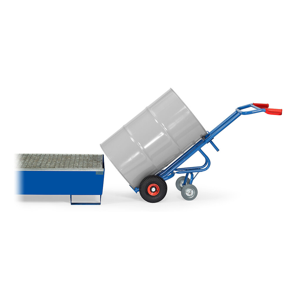 eine blaue FETRA® Karre aus Stahlrohr, mit zwei Rädern mit Luftbereifung auf roten Felgen, roten Handgriffen und geschwungener Stütze für Schräglage in Ruheposition mit zusätzlicher zweiter Achse mit zwei Stützradern, beladen mit einem grauen 200 Liter Fass aus Metall, vor einer Ölwanne mit Gitterrost, freigestellt auf weißem Hintergrund