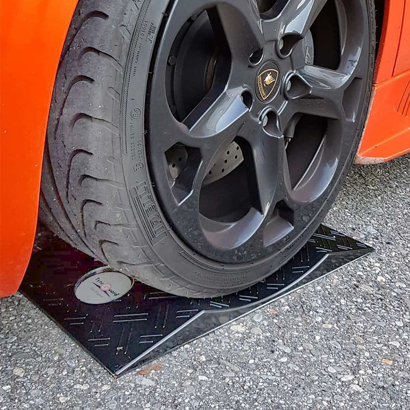 ein schwarzer konkaver TyreGuard® Reifenschoner aus hochfestem Kunststoff mit Antirutschmatte unter einem orangefarbenen modernen Sportwagen