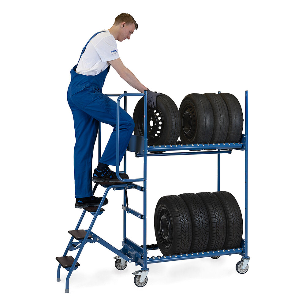 ein Arbeiter steht auf der Ausklappleiter eines blauen, mit 8 PKW-Reifen auf Stahlfelgen beladenen FETRA® Reifenkommissionierwagens aus Stahlrohr mit 4 Lenkrollen, davon 2 feststellbar, auf weißem Hintergrund
