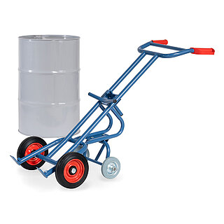 eine blaue FETRA® Karre aus Stahlrohr für den Transport von Fässern, mit zwei Rädern mit Vollgummi-Bereifung auf roten Felgen, roten Handgriffen und geschwungener Stütze für Schräglage in Ruheposition mit zusätzlicher zweiter Achse mit zwei Stützradern, dahinter ein graues 200 Liter Fass aus Metall, freigestellt auf weißem Hintergrund