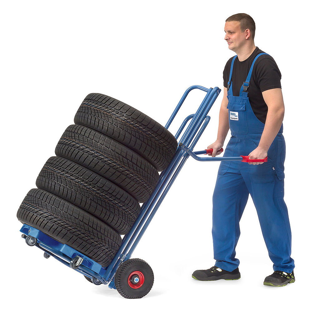 ein Arbeiter mit einer nach hinten gekippten blauen FETRA® Reifenkarre aus Stahlrohr mit Luftbereifung, darauf ein Reifenroller mit 4 PKW-Reifen, auf weißem Hintergrund