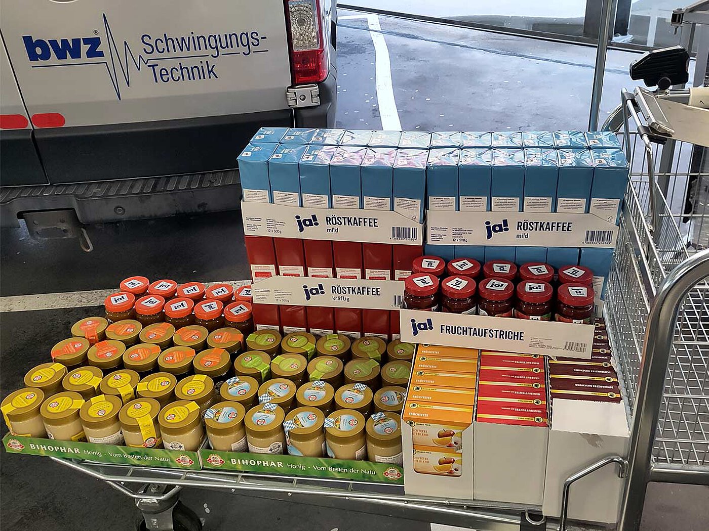 a C&C cart, loaded with trays of various packed food items, such as honey, tea, jams and filter coffee, in front of a silver-grey delivery van with the blue logo and script of company bwz Schwingungstechnik 