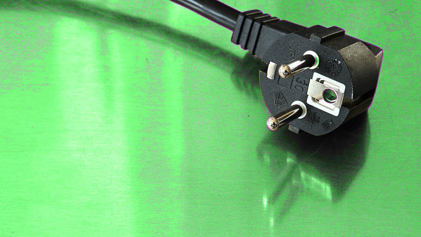 ein schwarzer Schuko-Stromstecker auf einer grünlich reflektierenden metallischen Oberfläche